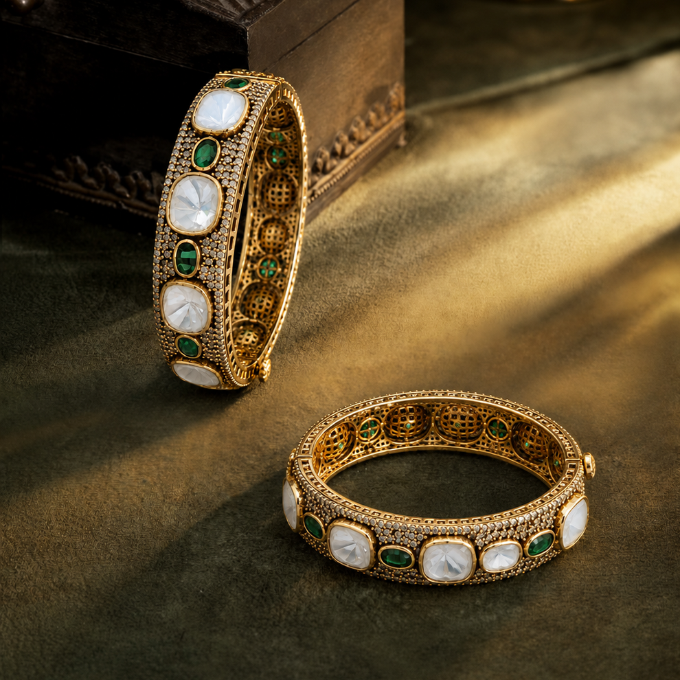 Two ornate gold bangles with white shell inlays and green stone accents, styled on a dark textured surface with a wooden box in the background.